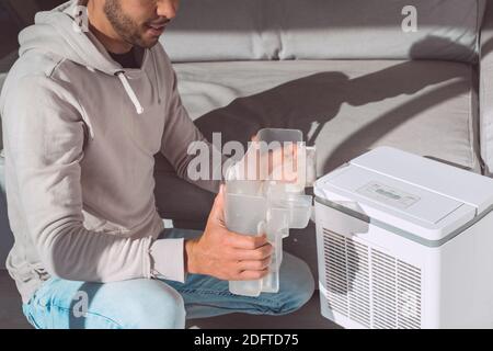 Man changing water container in air dryer, dehumidifier, humidity indicator. Humid air at home. Stock Photo