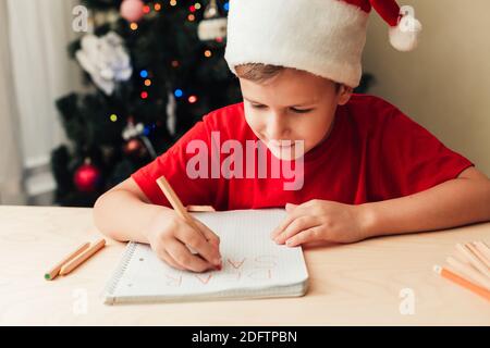 Smiling child boy wearing red hat write letter to Santa sitting by desk, Christmas tree on background, home interior Stock Photo