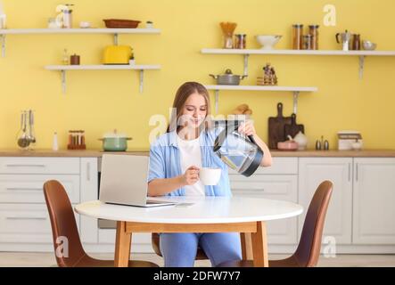 Young woman pouring boiled water from electric kettle into cup in kitchen Stock Photo