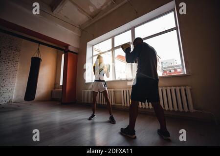 A man with boxing paws on his hands teaches the technique of hitting a novice girl in a light gym Stock Photo