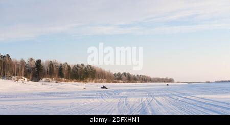 winter frozen river and forest on a river bank. man riding a snowmobile and dog running. people walking on a horizon. sunny day on the winter river. Stock Photo