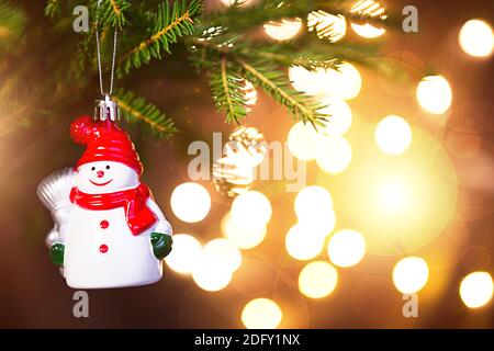 Christmas toy Snowman in a red hat with a scarf on live branch of a fir tree with Golden lights of garlands in defocus. New year, Christmas, holiday b Stock Photo