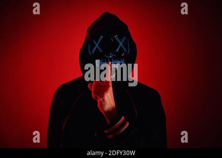Anonymous man in a hoodie hiding face behind neon glow scary mask on red background. Horror concept Stock Photo