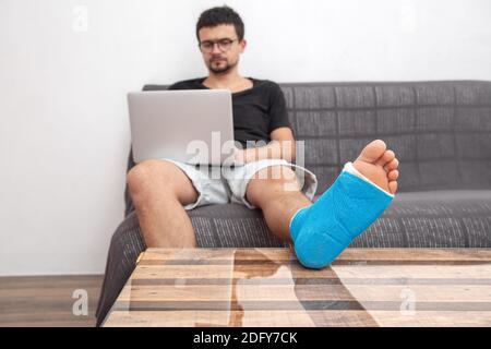 Man with glasses with broken leg in blue splint for treatment of injuries from ankle sprain working on a laptop on couch at home. Stock Photo