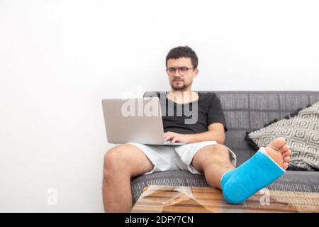 Man with glasses with broken leg in blue splint for treatment of injuries from ankle sprain working on a laptop on couch at home. Stock Photo