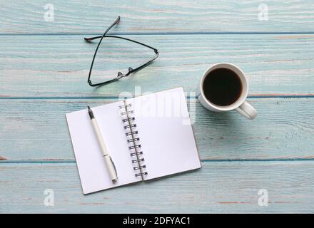 Open notepad with pen, spectacles and coffee on blue wood table. Top view. Stock Photo