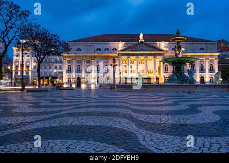 Low angle shot of fountain and illuminated facade of National Theatre as people walking at Rossio Square in Lisbon, Portugal Stock Photo