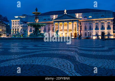 Low angle shot of fountain and illuminated facade of National Theatre as people walking at Rossio Square in Lisbon, Portugal Stock Photo