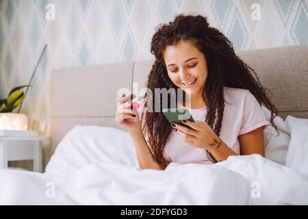 Positive young afro haired woman shop online using her smartphone sitting at home in bed paying with bank card Stock Photo