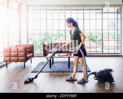 Young beautiful Asian woman, housewife wearing casual clothes using a vacuum cleaner to clean floor and carpet in living room near brown leather sofa Stock Photo