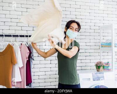 Young beautiful Asian woman, housewife wearing casual cloth and protective face mask shaking and drying shirt after washing before hanging on clothesl Stock Photo