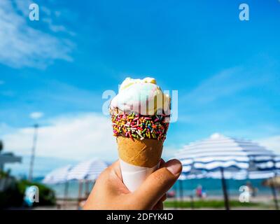 Ice cream in hand on beach and blue sky background. Rainbow color ice cream in cone with chocolate glaze with colorful sprinkles sugar topping. Stock Photo