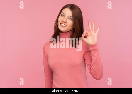 Everything is fine. Cheerful brunette woman in pink sweater showing ok gesture with hand looking at camera with toothy smile, approval. Indoor studio Stock Photo