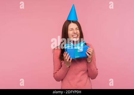 Extremely happy brunette woman in party hat holding blue present sincerely enjoying, receiving congratulations and gifts. Indoor studio shot isolated Stock Photo