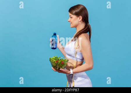 Profile portrait happy healthy slim woman in white sportswear holding bottle with mineral water and bowl with vegetable salad, healthy nutrition. Indo Stock Photo