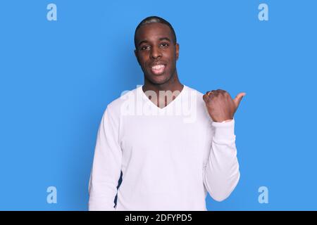 Black smiling Guy Pointing Thumb Aside At Blank Space For Text Standing On Blue Studio Background. Stock Photo