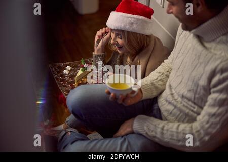 Charming woman in Santa hat and sweater holding tasty cake in hand near handsome man Stock Photo