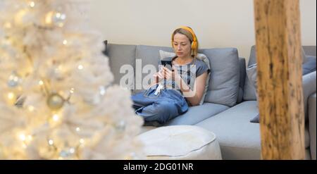Young thoughtful woman sitting indoors at home living room sofa using social media on phone for video chatting and staying connected with her loved Stock Photo