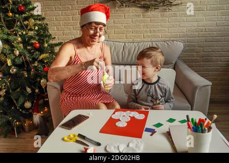 A Little boy with grandmother make Christmas craft, greeting card. Christmas tree and decoration on background. Stock Photo