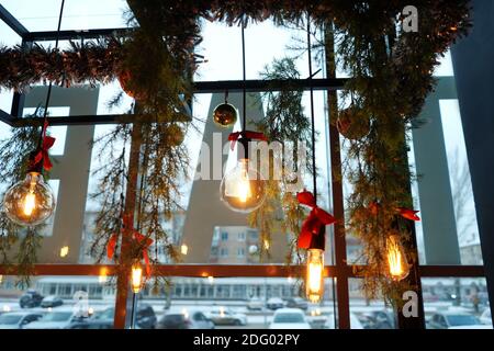 The long yellow light bulb hangs indoors in a cafe. Christmas design, decoration with fir branches and red bows. Window on background. Stock Photo