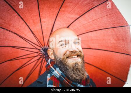 bald bearded man wearing a scarf and warm winter clothes holding a red umbrella as background.happy smiling young adult on white scenario in a studio Stock Photo