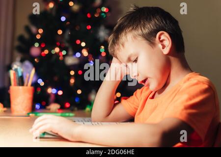 7 yeas old boy sitting at desk and reading book. Decorated Christmas tree on background Stock Photo