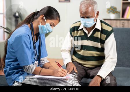 Young doctor or nurse writing prescription during home visiting to sick elder man while both worn face mask due to coronavirus covid-19 pandemic Stock Photo