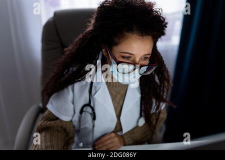 Portrait of young female afro haired doctor wearing glasses and protective face mask use her personal computer. Stock Photo
