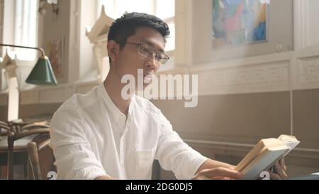 Young asian male student intently reading book in library of university. Attractive guy preparing for exams in campus Stock Photo