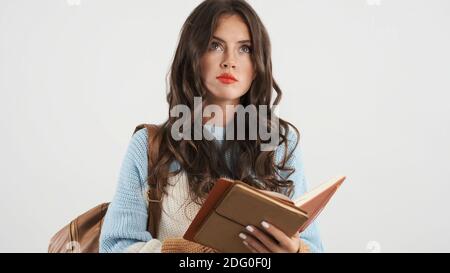 Beautiful pensive student girl with backpack thoughtfully reading textbook over white background Stock Photo