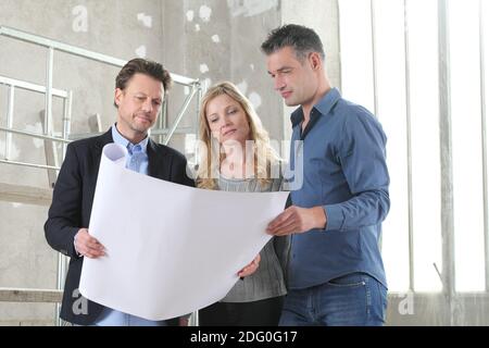 Architect showing house design plans to a young couple. House building goal concept. Meeting at interior construction site to talk about house appeara Stock Photo