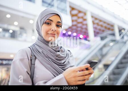Waist up portrait of smiling Middle-Eastern woman holding smartphone and looking at camera while enjoying shopping in mall, copy space Stock Photo