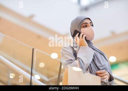 Portrait of modern Middle-Eastern woman wearing mask and speaking by smartphone while going down on escalator in shopping mall, copy space Stock Photo