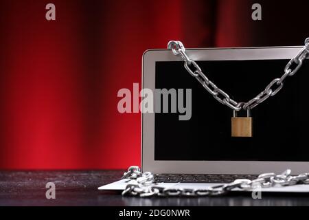 Laptop tied with an iron chain with a lock on a dark office table against black and red background. Cyber security and cyber crime. Stock Photo