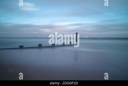 A long exposure captures streaky movement in the clouds and reduces the sea to a calm, smooth texture on the wet sand around the base of a groyne. Stock Photo