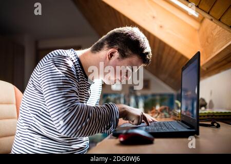 Down syndrome adult man sitting indoors in bedroom at home, using laptop. Stock Photo