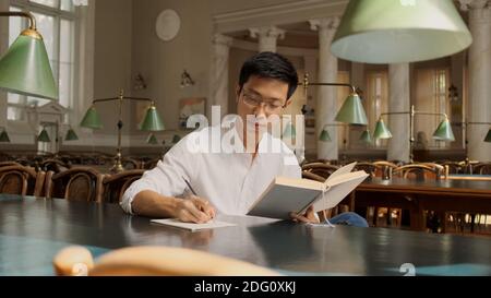Portrait of asian student with textbook intently making study notes in library. Young man studying in university Stock Photo