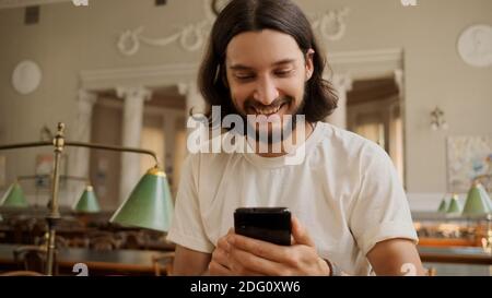 Portrait of cheerful casual student happily using smartphone in library. Attractive guy with phone studying in university Stock Photo