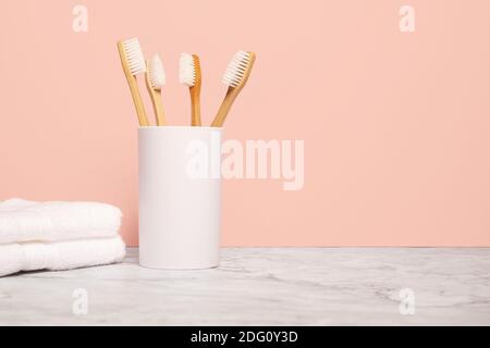Four natural wooden bamboo toothbrushes in a white cup next to a white cloth on a marble surface with pink wall with copy space and room for text Stock Photo