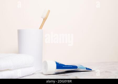 Single natural wooden bamboo toothbrush in a white cup next to a white cloth on a marble surface with tube of toothpaste copy space and room for text Stock Photo