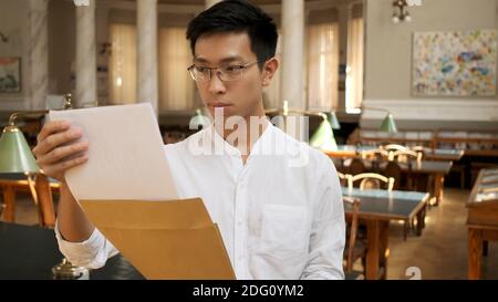 Asian male student intently opening envelope with exam results in university library Stock Photo
