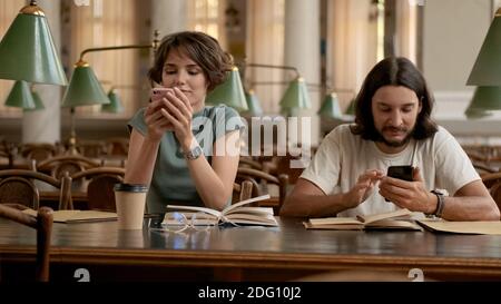 Two young students intently using smartphones while preparing for exams in library. Casual students studying together in university Stock Photo