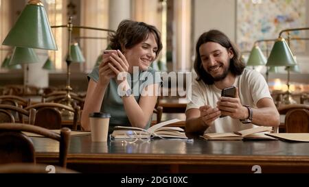 Attractive students happily using smartphones while preparing for exams in library. Casual smiling students studying together in university Stock Photo