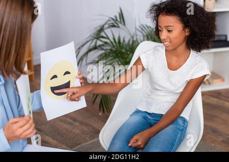 Smiling african american girl pointing with finger at happy expression on paper near psychologist on blurred foreground Stock Photo