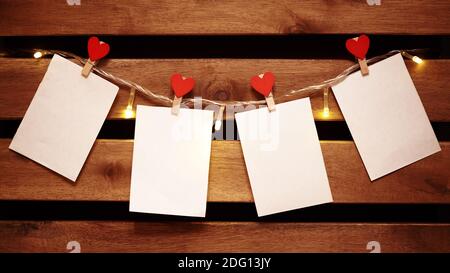 Blank White Piece of Paper hanging on Light Garland with Decorative Love Pins. Valentines Day Background with Copy Space. Cozy Wooden Background Stock Photo