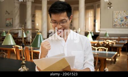 Portrait of asian student joyfully opening envelope with exam results in university library. Young attractive guy rejoicing exam results in campus Stock Photo