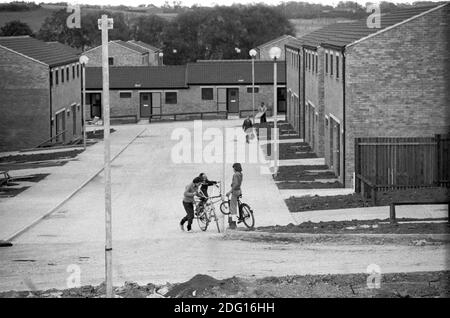 1970s England new town under development, houses built, but not all building works yet finished, gardens not planted, but new residents have  and are moving in.  Children playing in the empty street on their bikes 1977 Milton Keynes, Buckinghamshire  UK HOMER SYKES Stock Photo