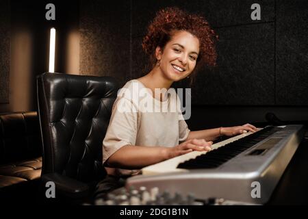 Young attractive woman happily looking in camera playing on electric piano in recording studio. Female musician working in studio Stock Photo