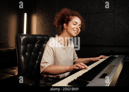 Young pretty woman sensually playing on electric piano and singing a song in recording studio. Attractive musician working in studio Stock Photo