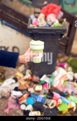 Female hand holding reusable eco cup and a lot of garbage on the background Stock Photo